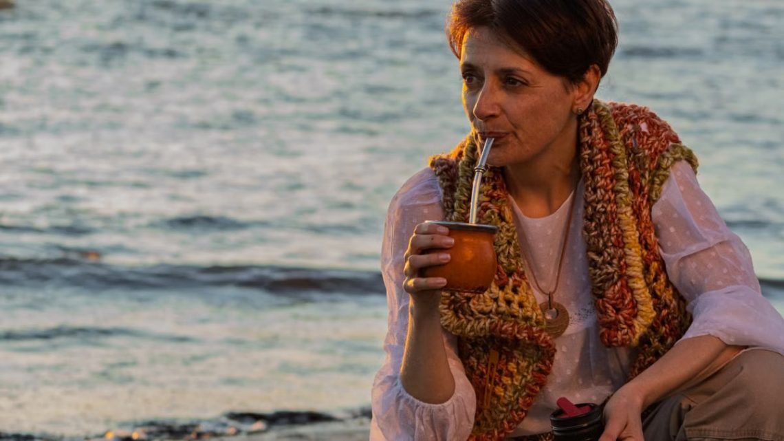 A woman sipping yerba mate while relaxing on a Montevideo beach during sunset.