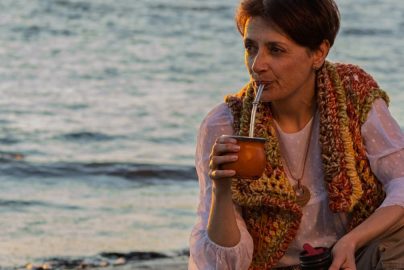 A woman sipping yerba mate while relaxing on a Montevideo beach during sunset.